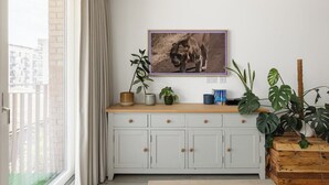 Een kamer met een wit kastje, een lijst met een foto aan de muur, en een raam met gordijnen.