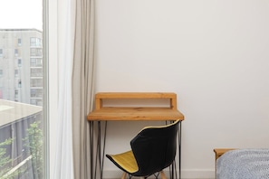 Ein Eckzimmer mit einem Holztisch, einem schwarzen Stuhl mit gelber Sitzfläche und einem Bett mit gemusterten Betttextilien.