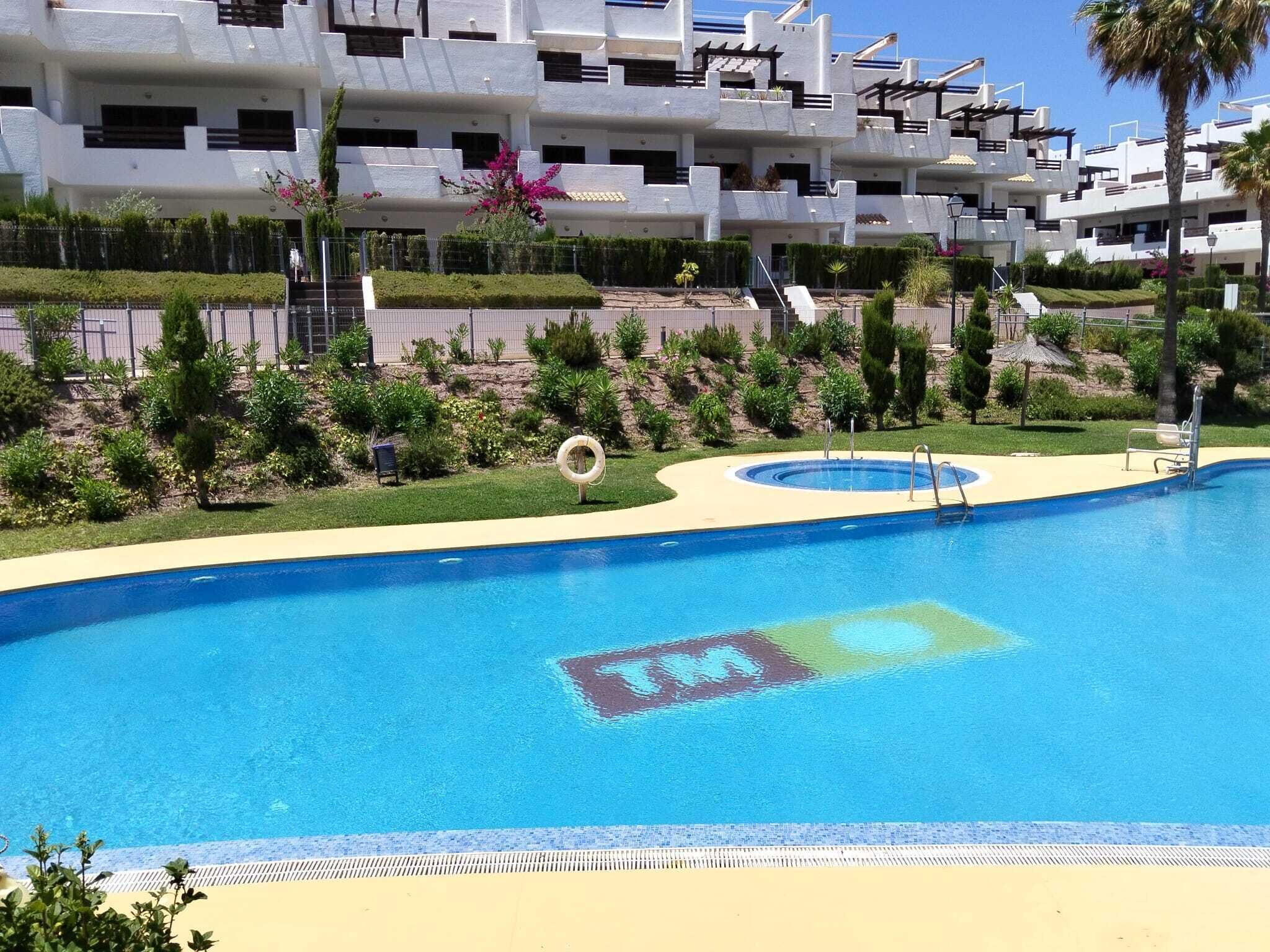 A swimming pool with a sunbathing area, surrounded by greenery and a modern building with balconies.