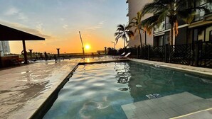 En swimmingpool med solnedgang i baggrunden, omgivet af palmer og liggestole.