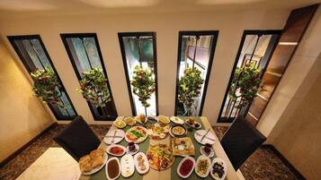 A dining table with various dishes, surrounded by chairs, in a room with large windows and potted plants.