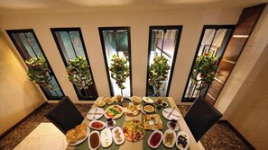 A dining table with various dishes, surrounded by chairs, in a room with large windows and potted plants. - Royal Berk Hotel (VAN)