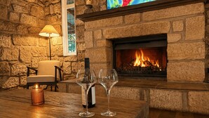 A stone fireplace with a lit fire, a wooden table with a wine bottle and glasses, a chair, and a wall-mounted TV. - Highwoods Cottage - Authentic charm in the heart of Wine Country (Pokolbin)