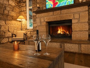 A stone fireplace with a lit fire, a wooden table with a wine bottle and glasses, a chair, and a wall-mounted TV. - Highwoods Cottage - Authentic charm in the heart of Wine Country (Pokolbin)