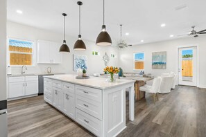 A modern kitchen with white cabinetry, a large island, pendant lighting, and a dining area with white chairs. - Wanderlust by AvantStay | Walk to Beach + Pool | New Construction (Panama City Beach)