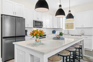 A modern kitchen with white cabinets, stainless steel appliances, and a central island with stools. - Wanderlust by AvantStay | Walk to Beach + Pool | New Construction (Panama City Beach)