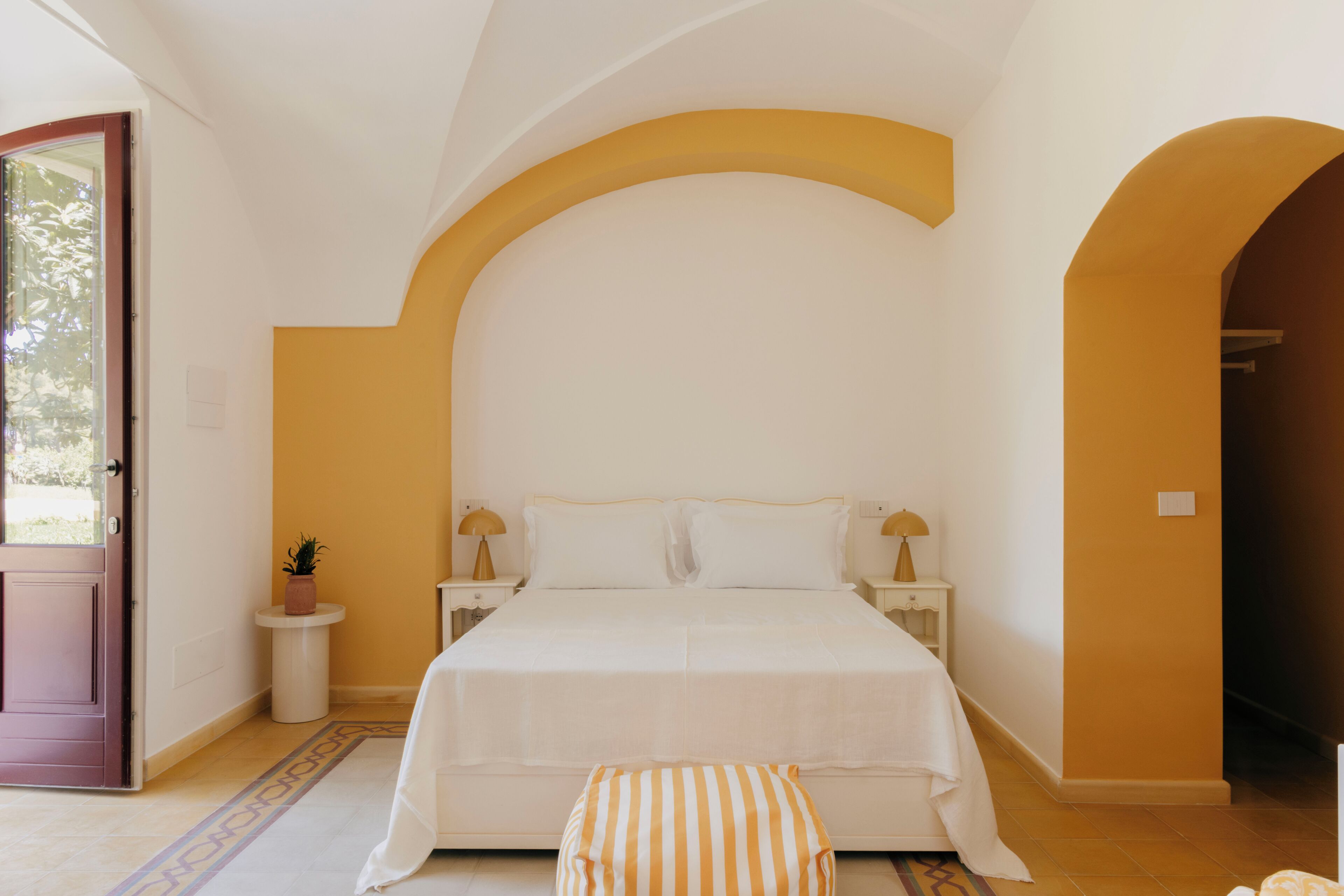 Une chambre à coucher avec un lit, des tables de chevet, une petite table, une plante en pot et une porte en arc de cercle.