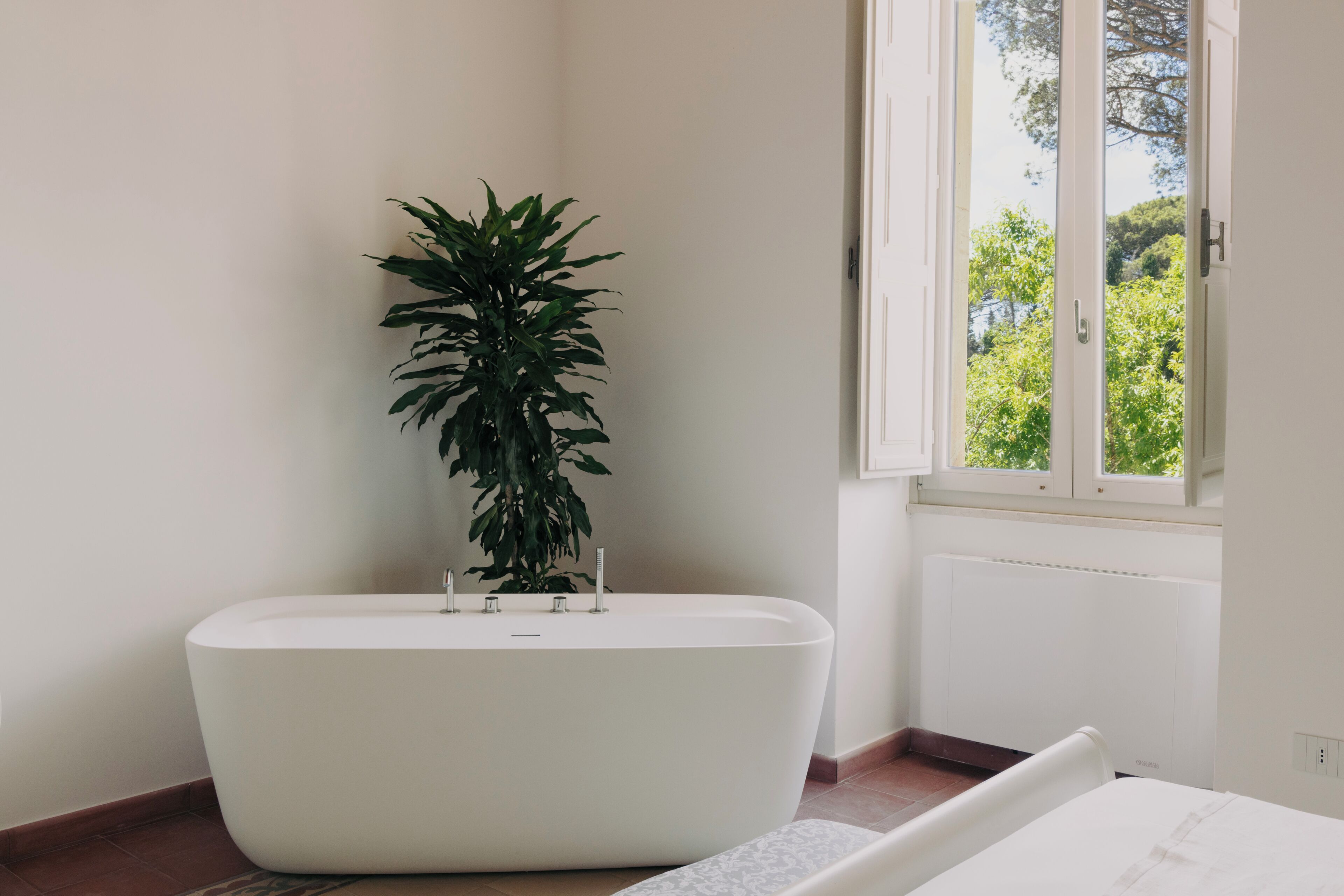 A modern bathroom with a freestanding bathtub, a potted plant, and a window with a view of greenery.