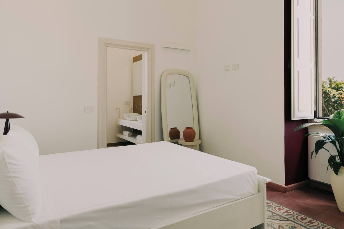 Un dormitorio con cama, un espejo, estante con artículos de tocador y ventana con vistas a zonas verdes.