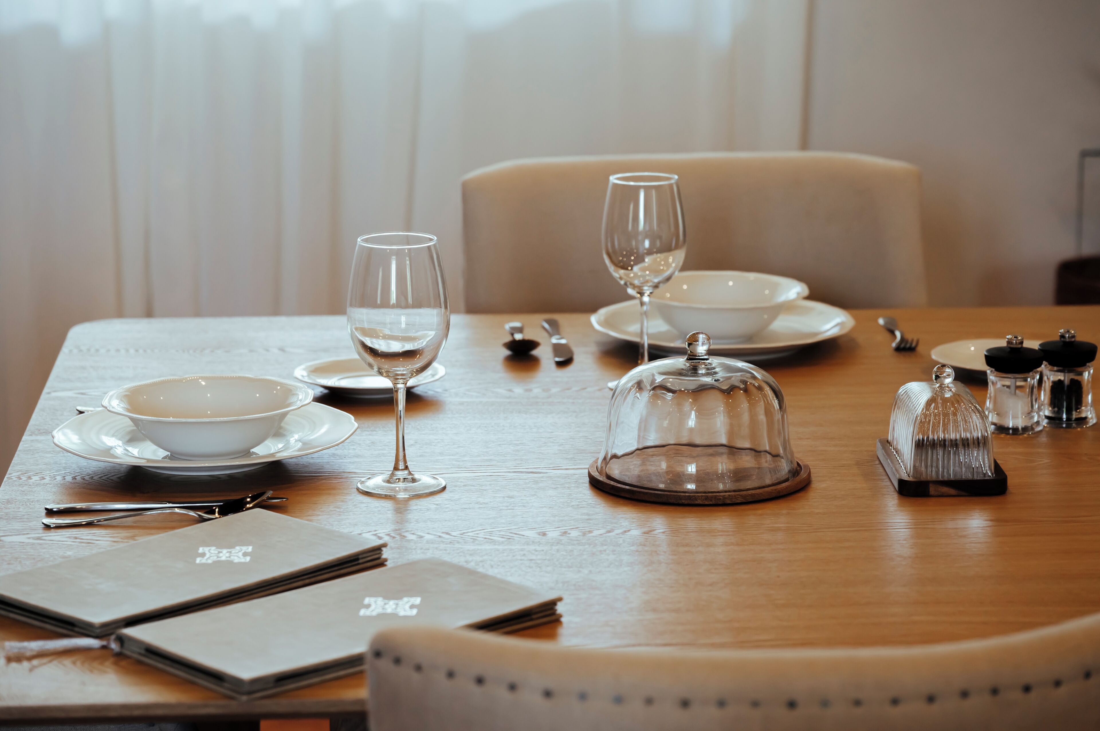 Una mesa de comedor dispuesta con platos, vasos, cubiertos, una campana de cristal como centro de mesa, saleros y pimenteros, y un menú.