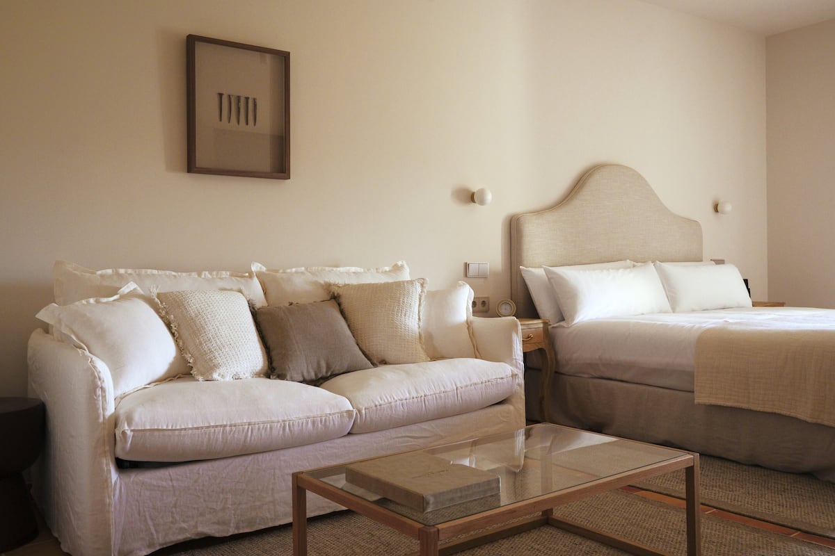 Une pièce avec un canapé beige, un lit doté d’une tête de lit, une table de chevet et un tableau encadré au mur.