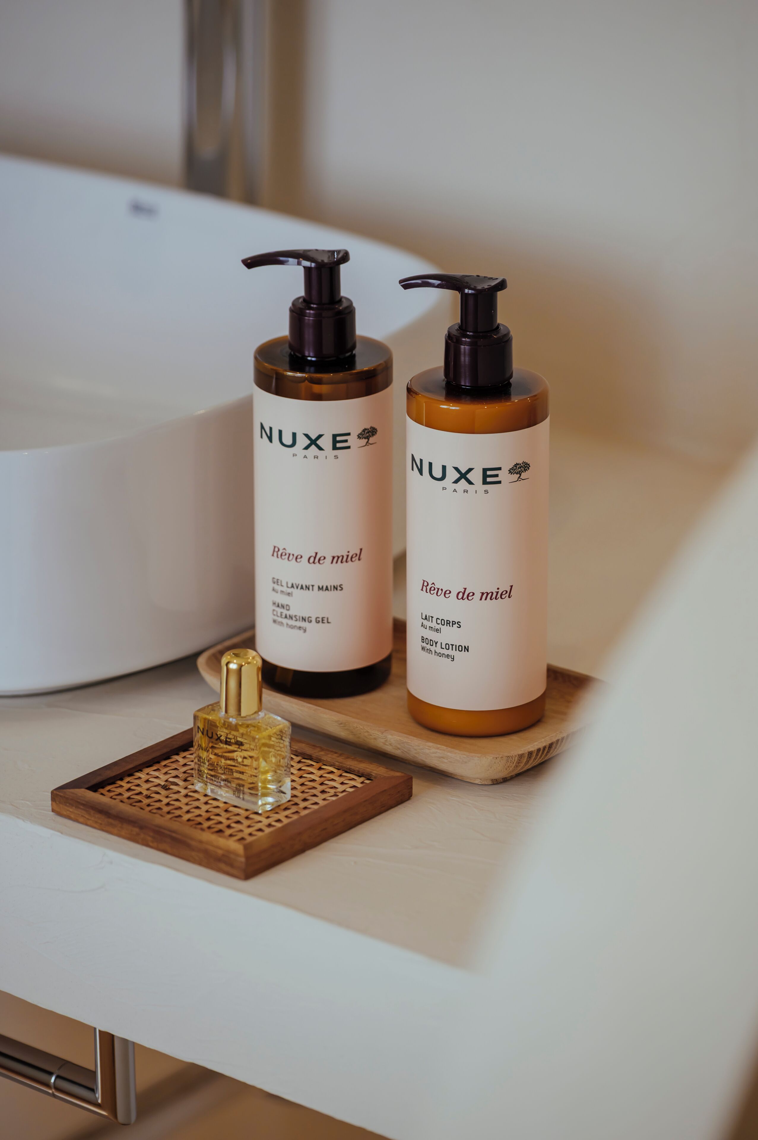Dos botellas de productos para el cuidado de la piel NUXE Paris sobre una bandeja de madera junto a un lavabo blanco.