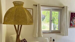 Quarto com uma cadeira branca, uma mesinha redonda decorada, um abajur e uma janela com cortinas.