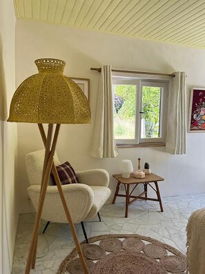 Ein Zimmer mit weißem Sessel, einem kleinen runden Tisch mit Dekoration, einer Stehlampe und einem Fenster mit Vorhängen.