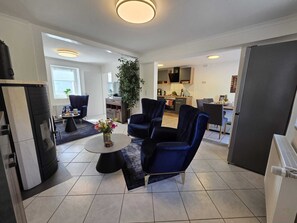 Un salon moderne avec une table basse ronde, deux fauteuils bleus, une plante en pot et une cuisine en arrière-plan.