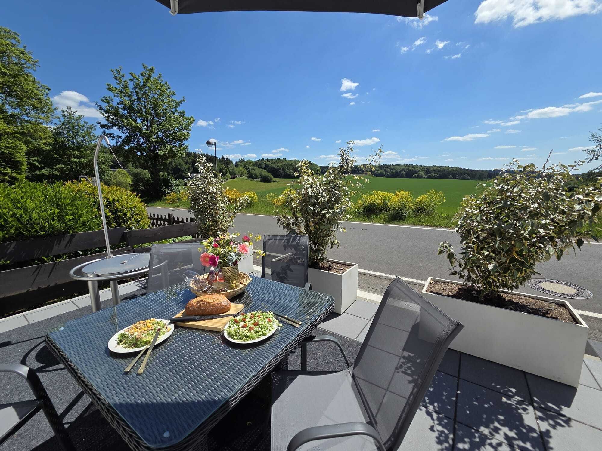 Un área de comedor al aire libre con una mesa dispuesta para dos personas, rodeada de sillas, y una vista de paisaje verde al fondo.