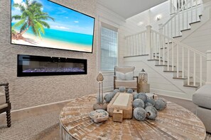 Ein Wohnzimmer mit großem Fernseher, der eine Strandansicht zeigt, einem Kamin, einer Treppe und einem runden Couchtisch mit Dekoration.