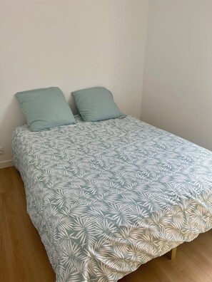 Una cama individual con una colcha estampada y dos almohadas, suelo de madera y pared lisa.