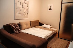 A bedroom with a brown sofa, a bed with patterned bedding, a framed abstract artwork, and a framed magazine on the wall.