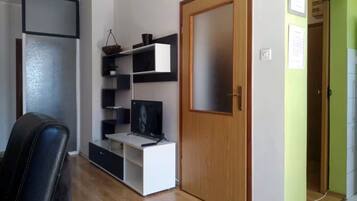 Une pièce avec une porte en bois, un téléviseur posé sur un meuble blanc et une chaise noire.