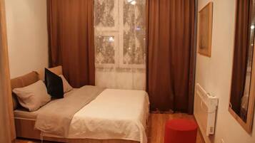 A small, cozy bedroom with a bed, a red ottoman, a window with curtains, and a radiator.