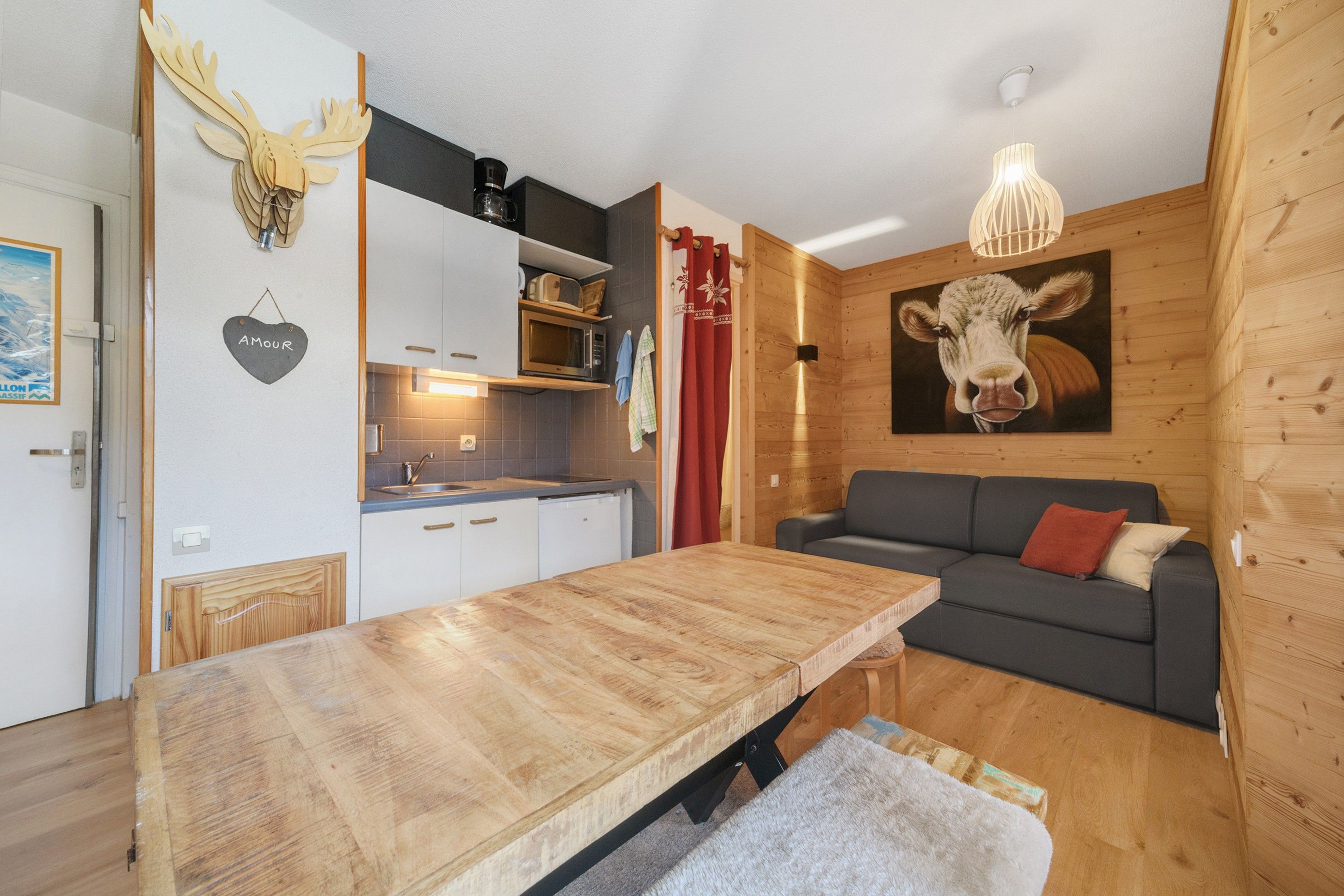 A cozy cabin interior with a wooden dining table, a sofa, a kitchen area, and a mounted cow painting.