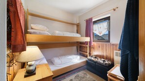 Een slaapkamer met stapelbed, een houten hoofdbord, een nachtkastje met lamp, een klein krukje en een raam met gordijnen.