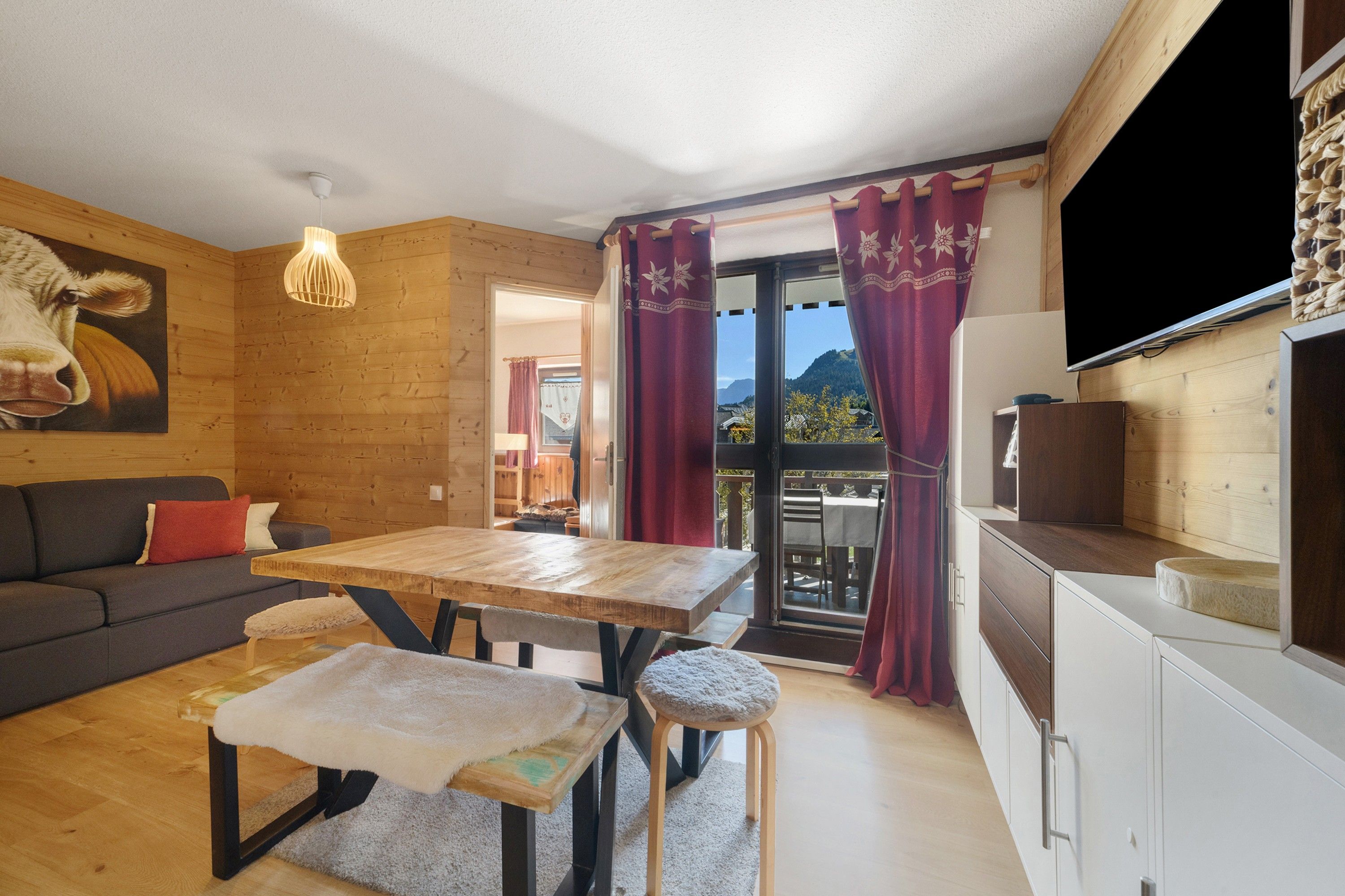 A cozy room with a wooden ceiling, a sofa, a table with stools, and a TV mounted on the wall.