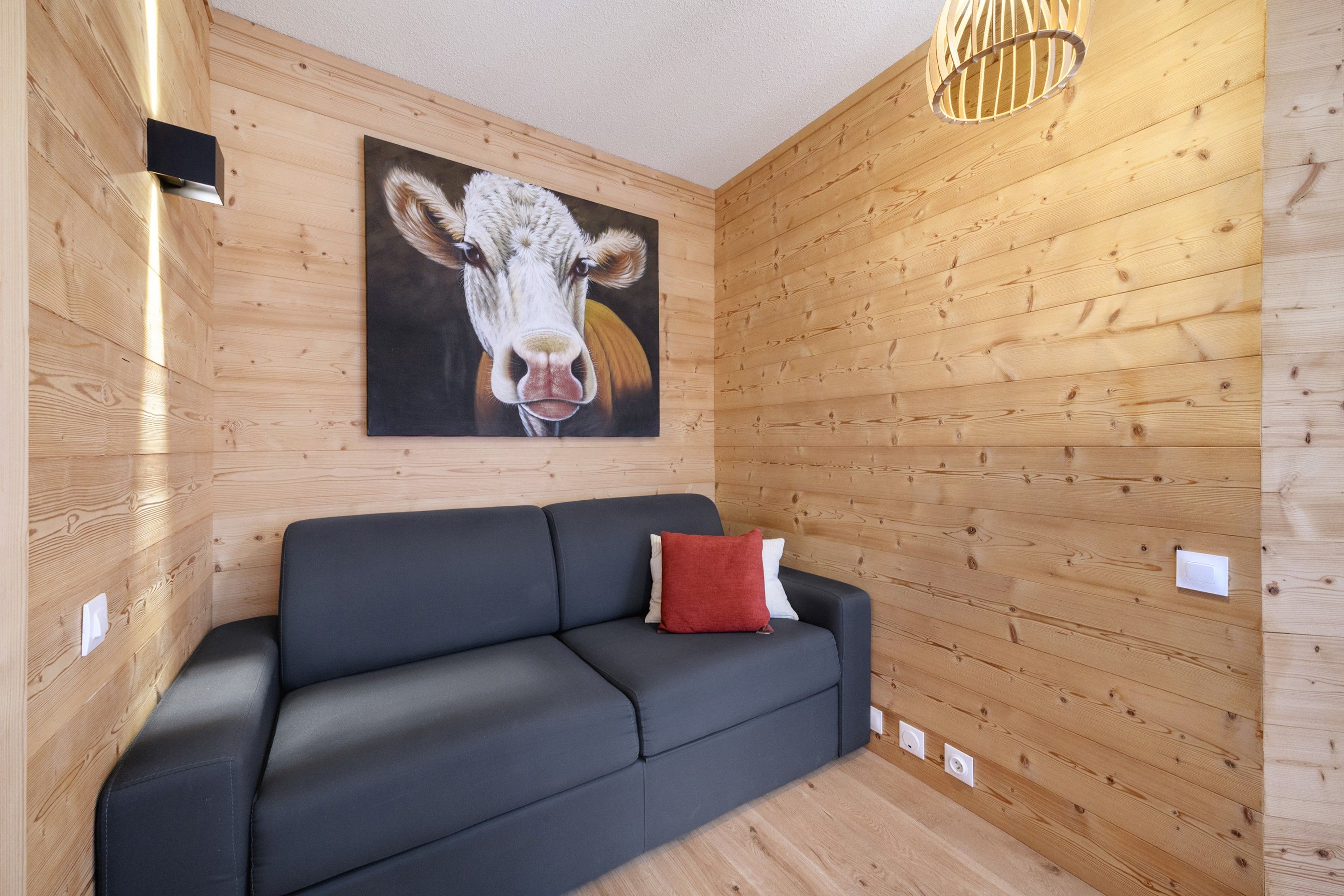Ein modernes Interieur mit einer Holzpaneelausstattung, einem schwarzen Sofa, einem roten Kissen und einem Gemälde mit einer Kuh.