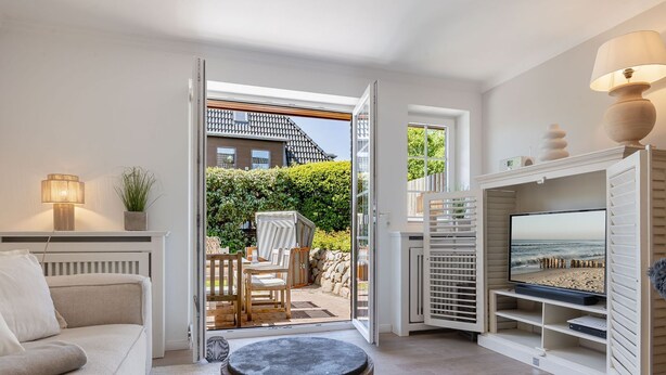 Ein Wohnzimmer mit einer Couch, einem kleinen runden Tisch, einem Fernseher auf einem Schrank und einer Glastür zu einem Balkon mit Gartenmöbeln.