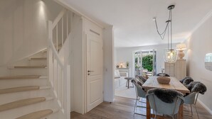 Ein modernes Interieur mit einer Treppe, einem Essbereich mit einem Holztisch und Stühlen sowie einem Gartenblick durch eine offene Tür.
