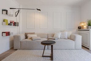A living room with a sofa, a coffee table, a floor lamp, a radiator, and a shelf with books and decorative items. - Beach Love Sylt, Germany (Sylt)