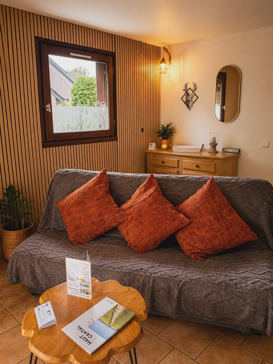 A room with a grey sofa, wooden panel walls, a wooden cabinet, a mirror, and a window with a view of greenery.