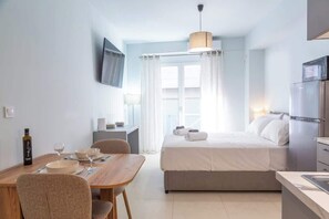 Ein kompaktes Hotelzimmer mit Bett, einer kleinen Küchenzeile, einem Essbereich mit Tisch und Stühlen, einem an der Wand montierten Fernseher und einem Fenster mit Vorhängen.