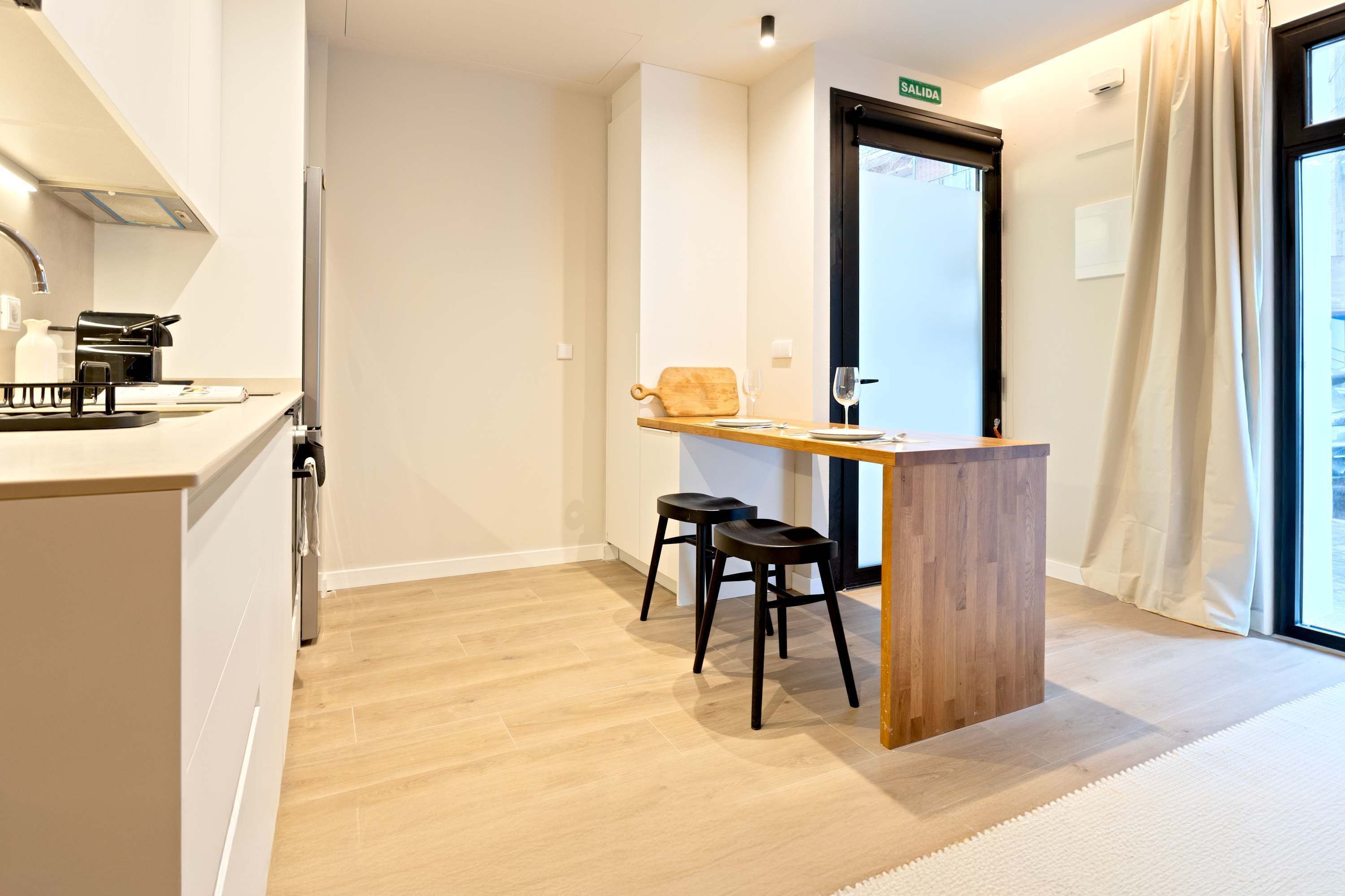 Una cocina moderna con cafetera, mostrador de madera, dos taburetes y una puerta que indica 'SALIDA'.