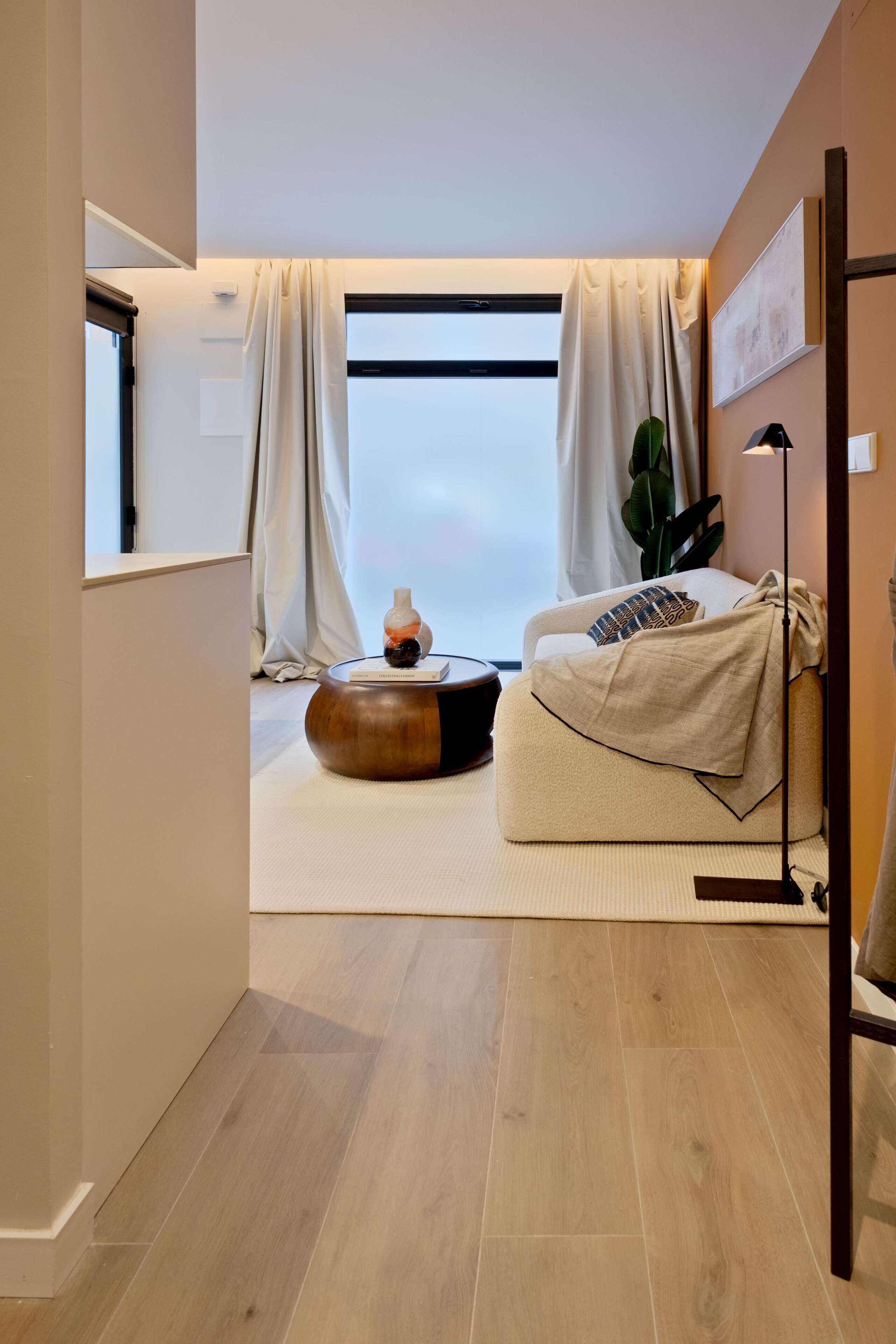 Una sala de estar moderna con un sofá, una mesa de centro redonda de madera y un ventanal con cortinas.