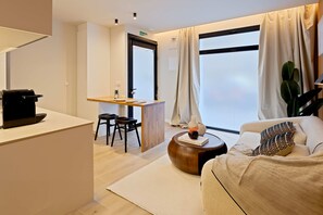 Una habitación de hotel moderna con escritorio, sofá, un ventanal con cortinas y una planta.