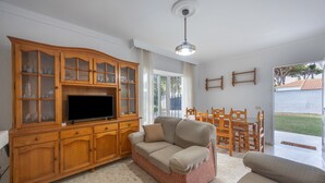 Ein Wohnzimmer mit Holzschrank, einer Couch, einem Fernseher und einem Essbereich mit Holztischen und einer Tischplatte.