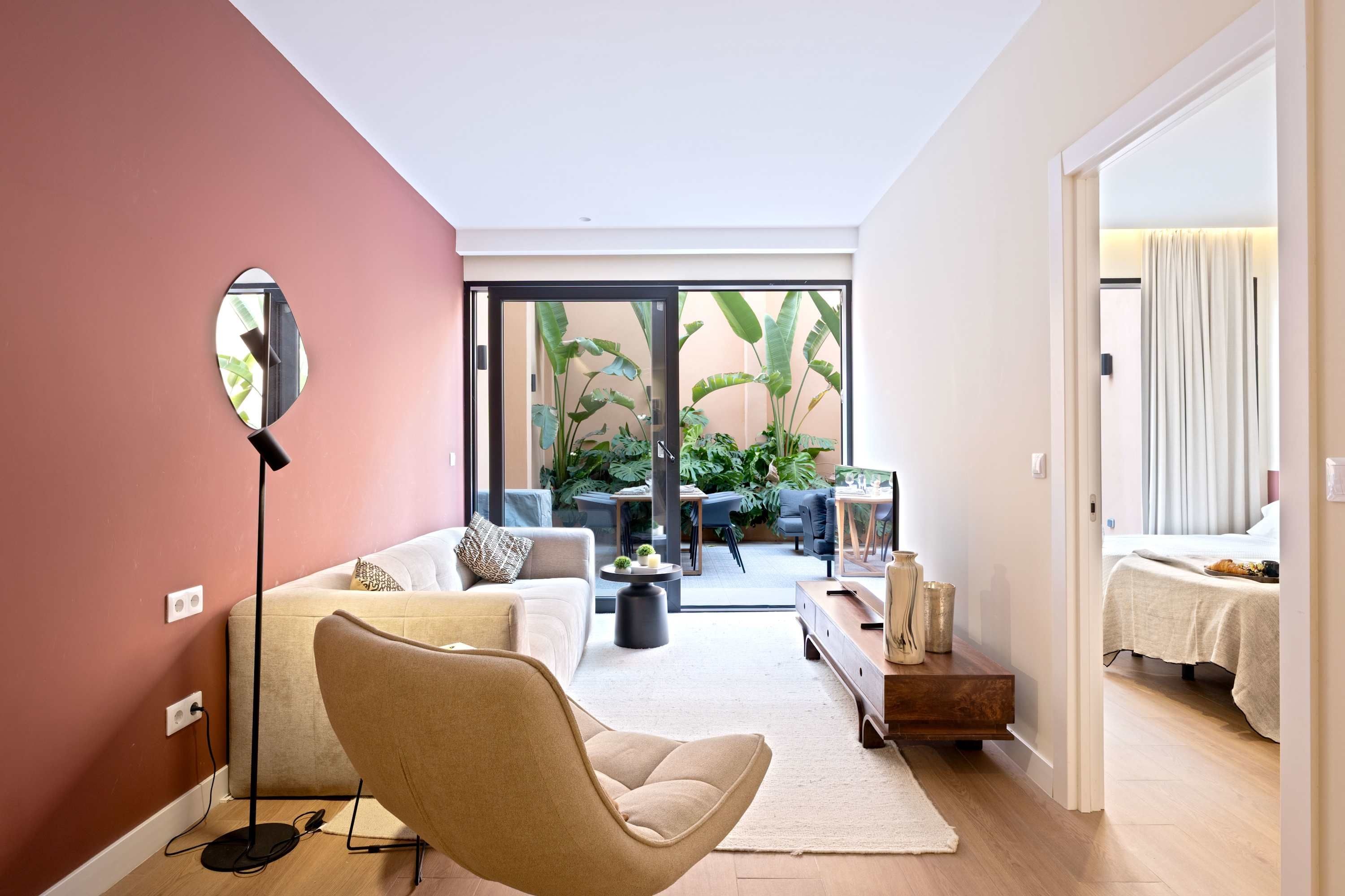Ein modernes Wohnzimmer mit einer Couch, einem Sessel und einem großen Spiegel. Es gibt einen Essbereich mit Tisch und Stühlen, und einen Blick auf einen Garten mit üppigen Pflanzen.