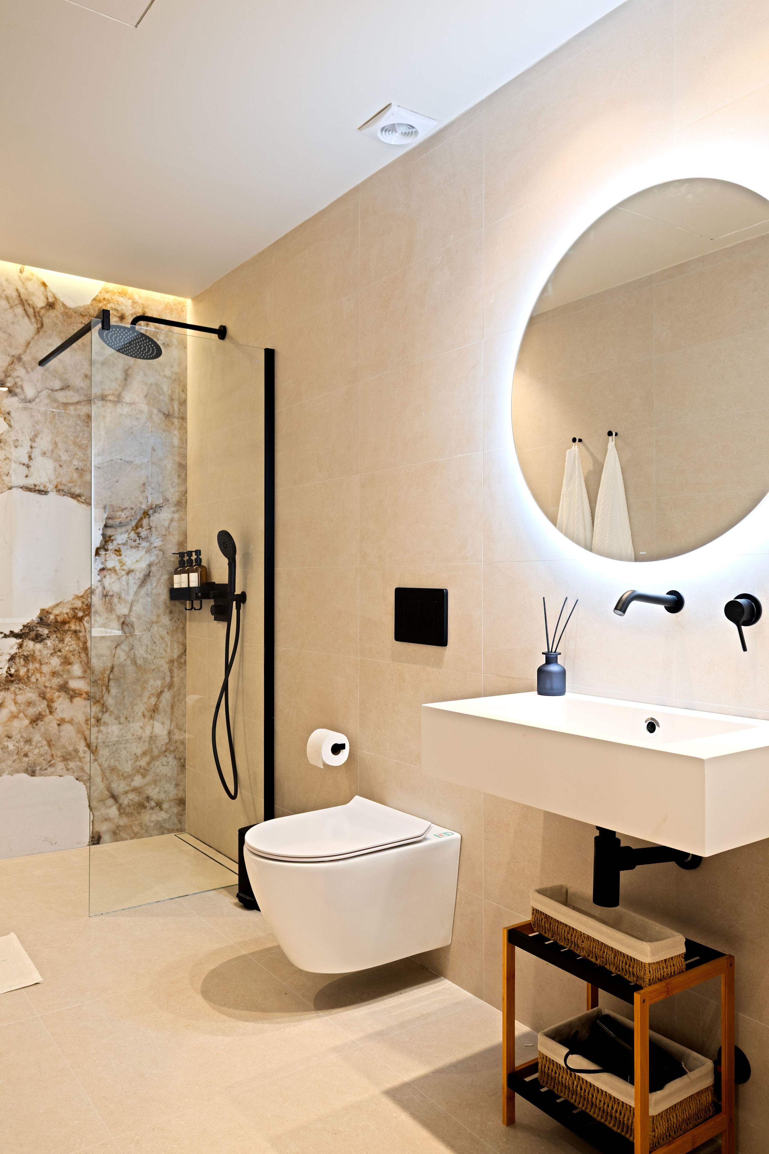 Een moderne badkamer met een marmeren wand, een wit toilet, een wastafel met spiegel en een douchegedeelte.