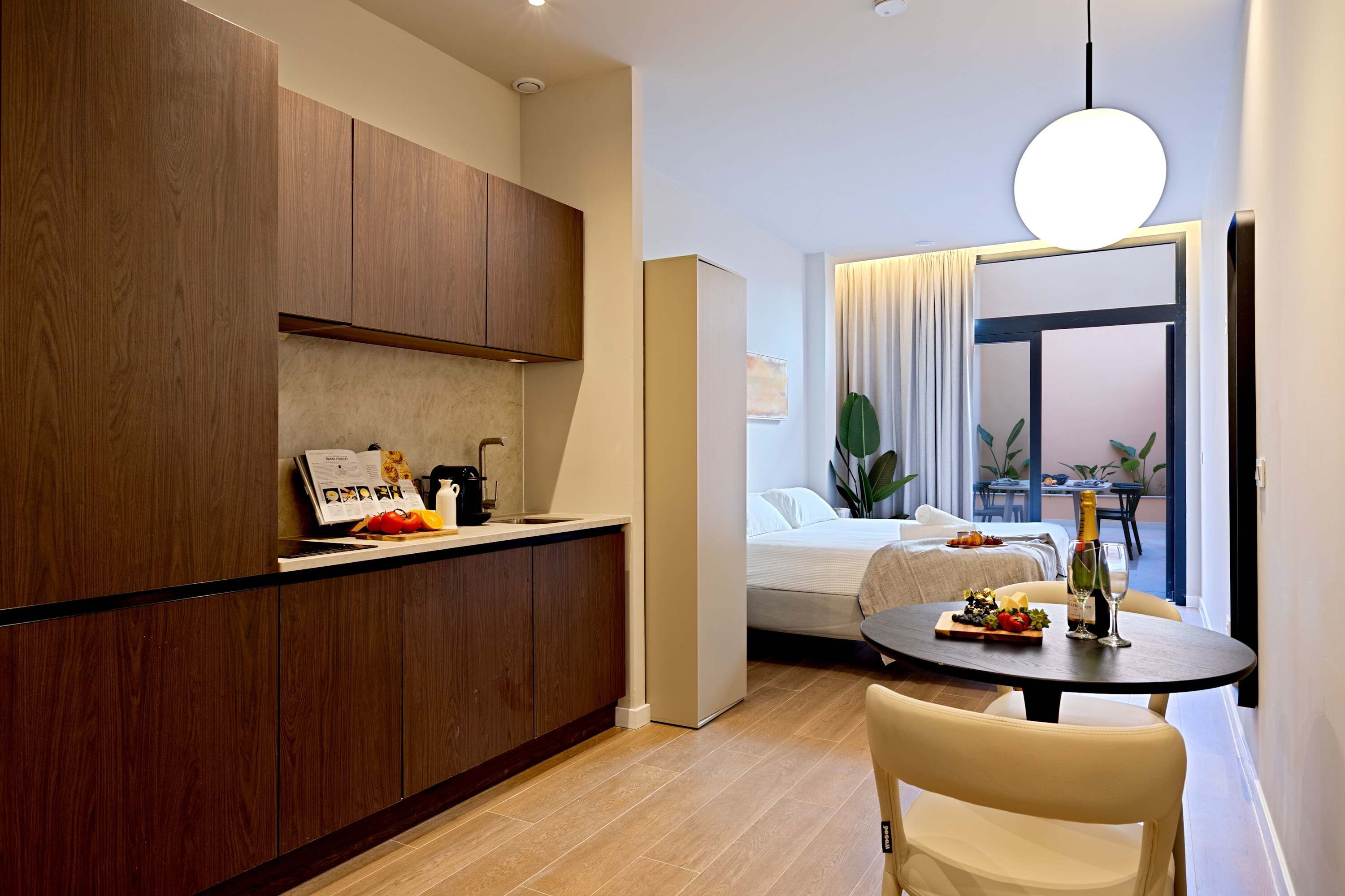 A modern kitchen with wooden cabinets, a dining area with a table and chairs, and a bedroom with a bed and curtains.