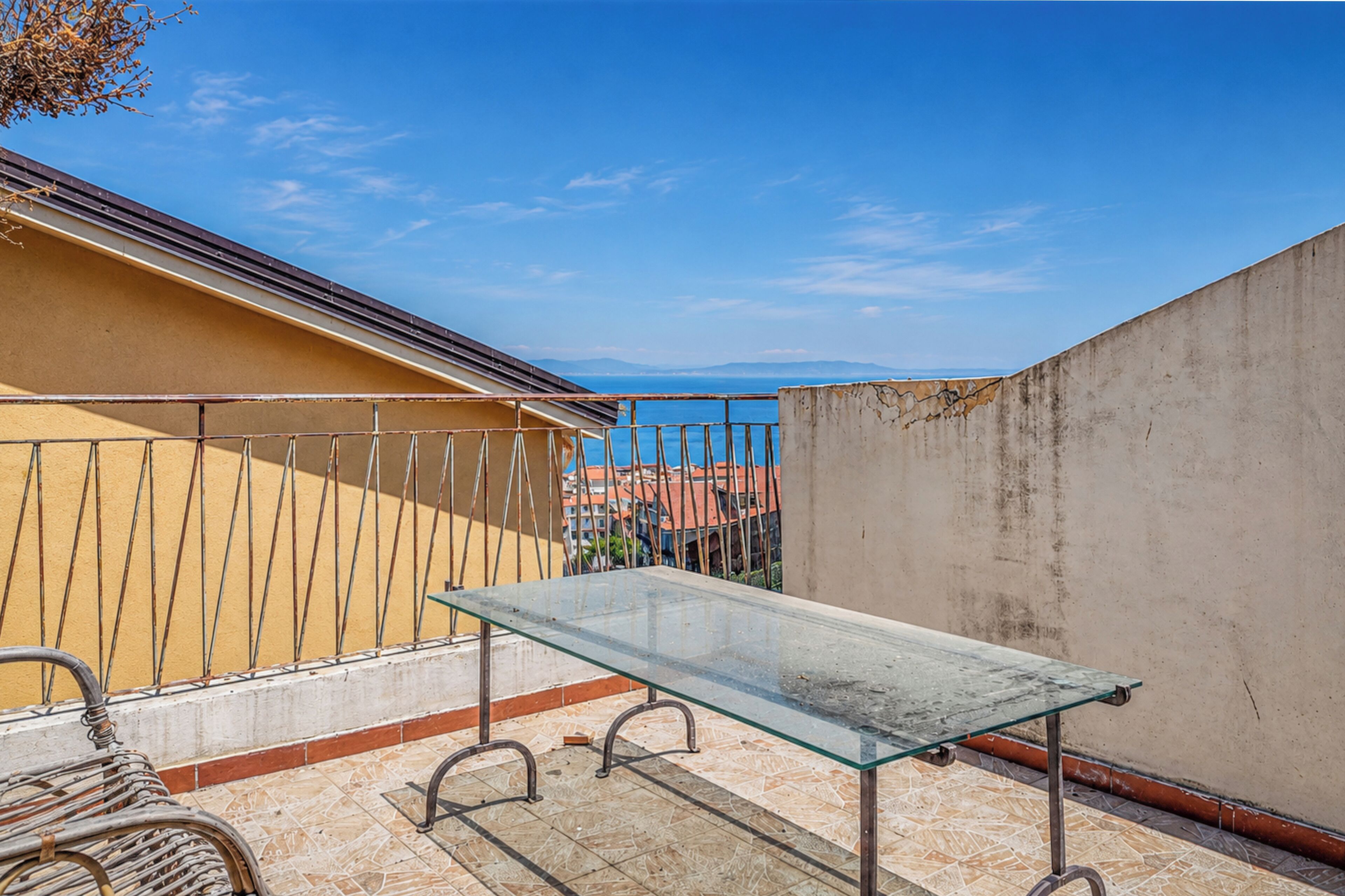 Un balcón con una mesa de vidrio, barandilla de metal y vistas al mar y edificios.