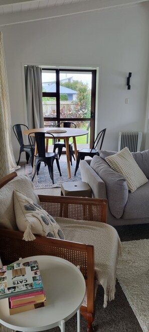 A living room with a sofa, a coffee table with books, a dining area with chairs and a table, and a view through a window. - Point Wells Village home with large covered deck. Pet friendly! (Point Wells)