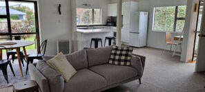 A living room with a sofa, a dining table with chairs, a kitchen area with a refrigerator, and a view through a large window.