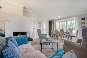 A living room with a fireplace, a dining area with a table and chairs, and a view of the outdoors through large windows. - Wääfblik Seestraße - Wääfblik – Your vacation home on Sylt with a sauna and ocean view (Wenningstedt-Braderup (Sylt))