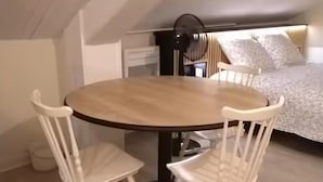 Uma pequena mesa redonda para refeições com duas cadeiras brancas, um ventilador de teto e uma cama com roupa de cama estampada ao fundo.