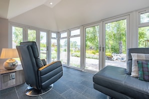 A room with a massage chair, a sofa with a plaid pillow, and a lamp on a side table.