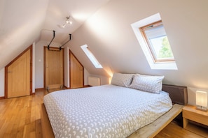 A bedroom with a sloped ceiling, a skylight, a bed with patterned bedding, wooden flooring, and a nightstand with a lamp.