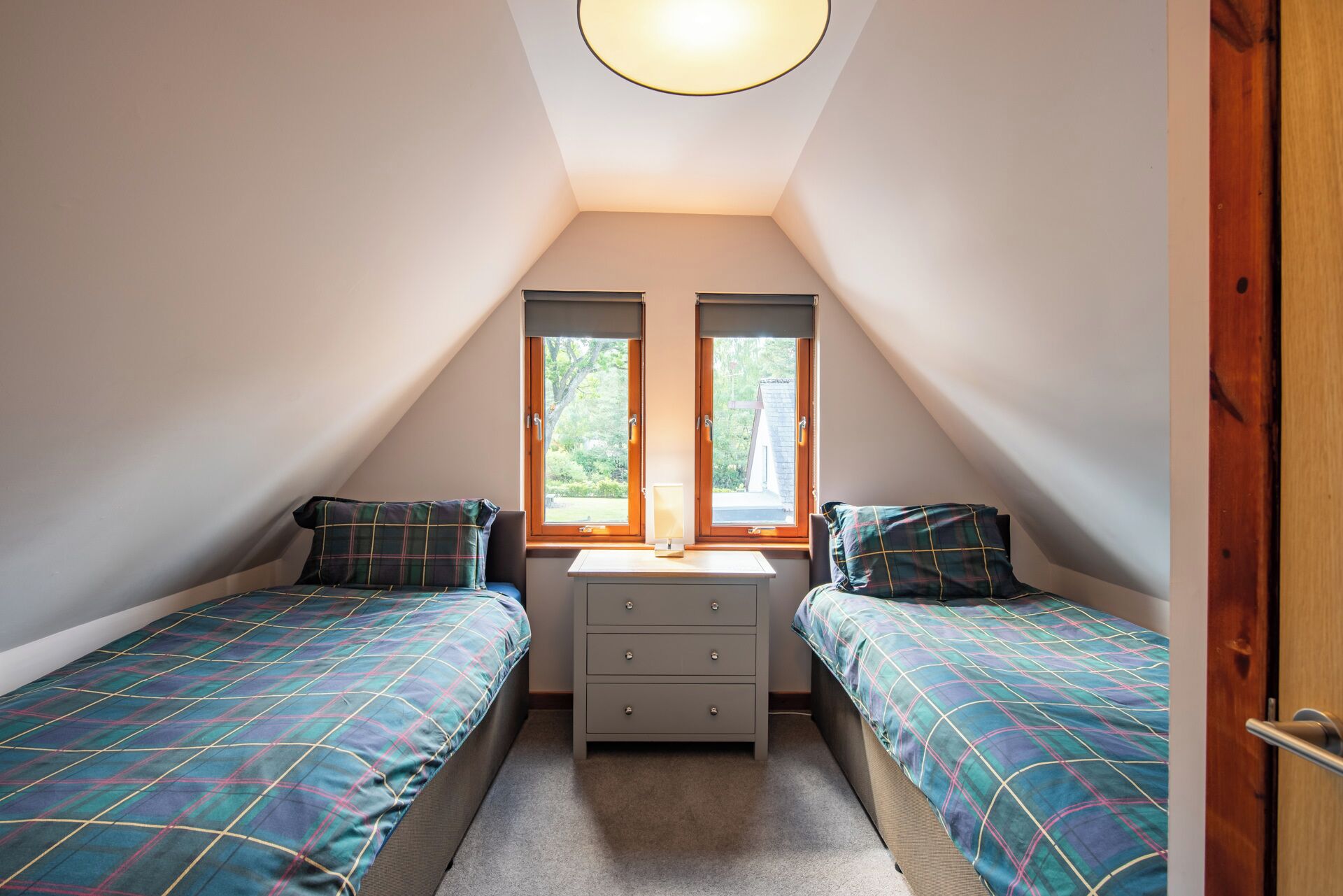 Een kleine, driehoekige slaapkamer op zolder met twee eenpersoonsbedden, een nachtkastje in het midden en twee ramen.