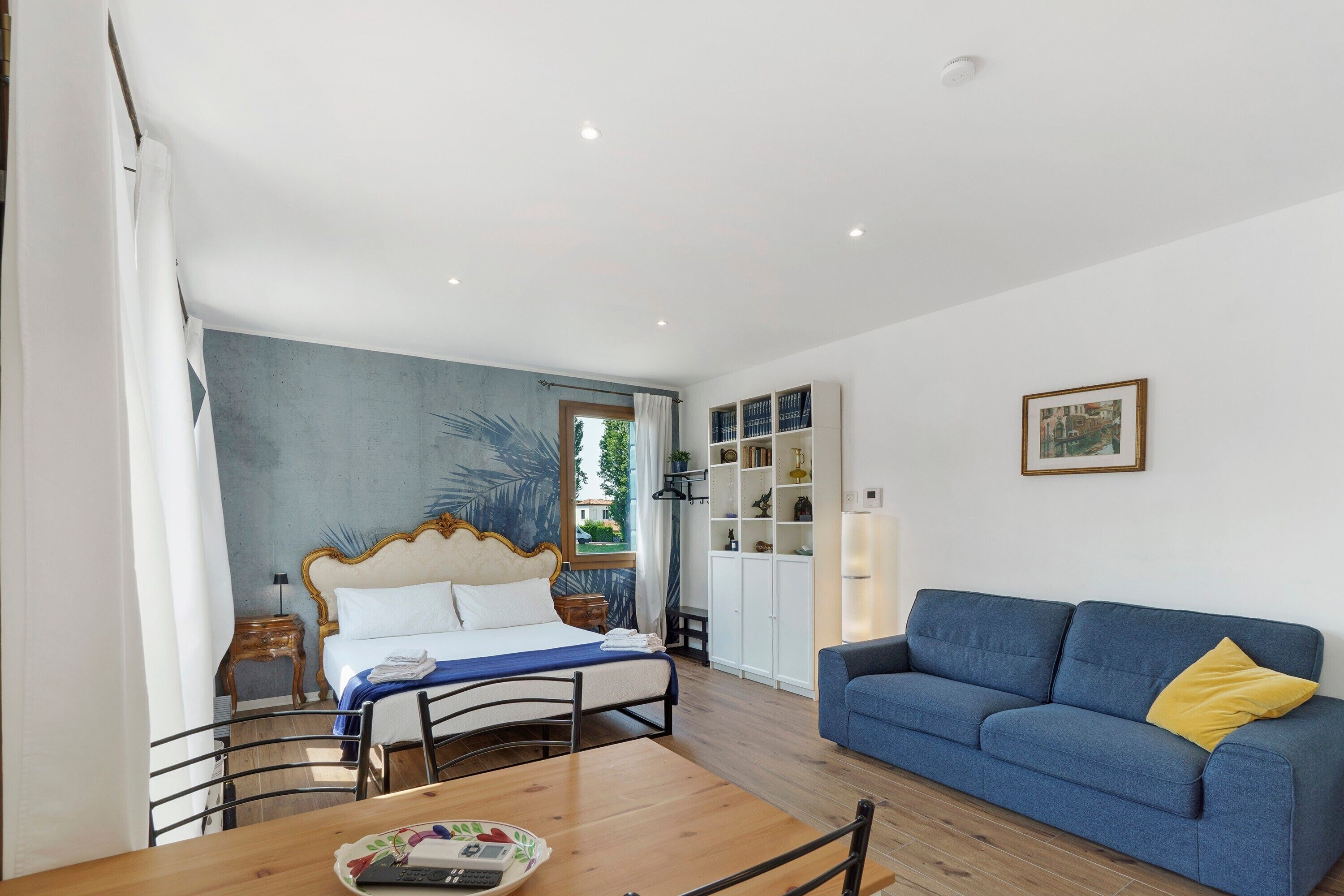 A bedroom with a bed, a blue sofa, a dining table with chairs, a bookshelf, and a window.